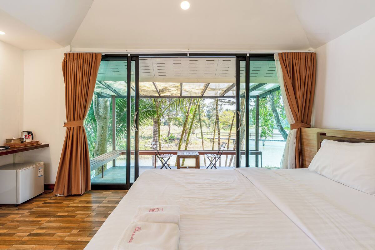A hotel room with a large bed, a desk with a computer, and a view of palm trees through a glass door.