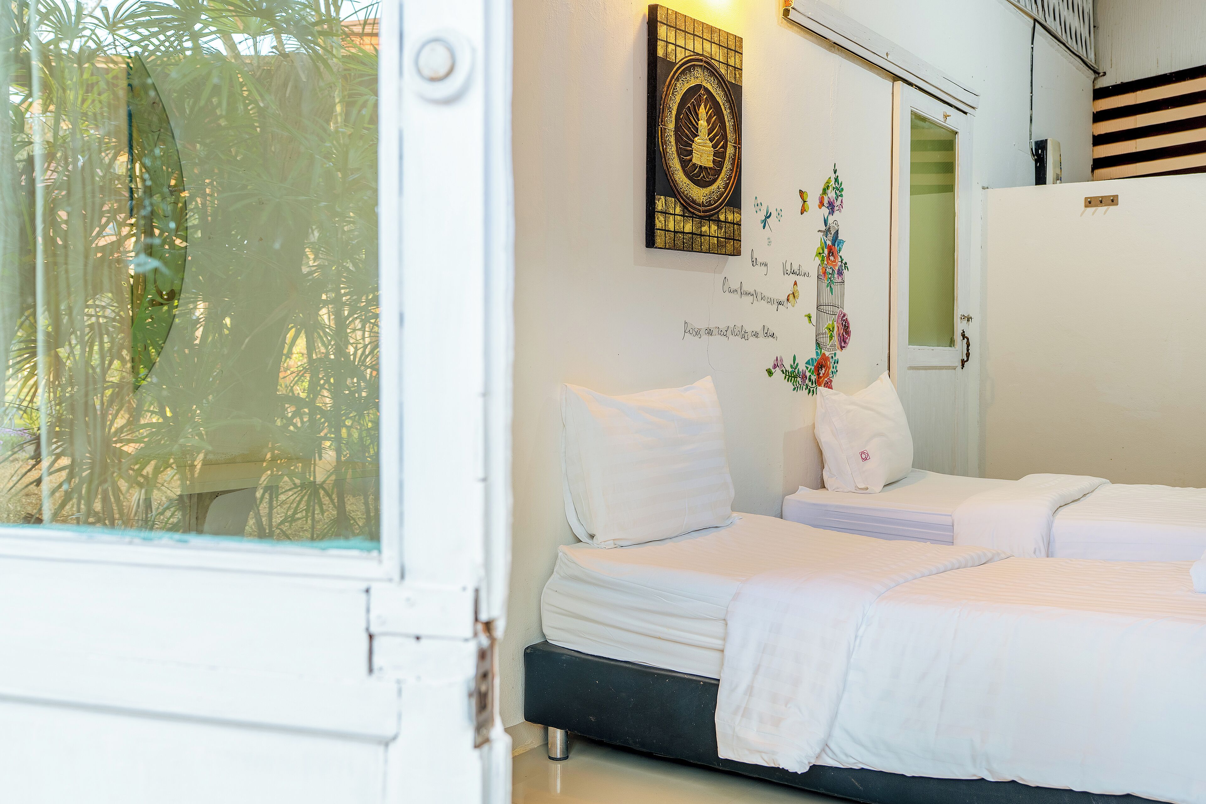 Un dormitorio con dos camas, una ventana con vistas a la vegetación, un adorno en la pared y una puerta blanca.