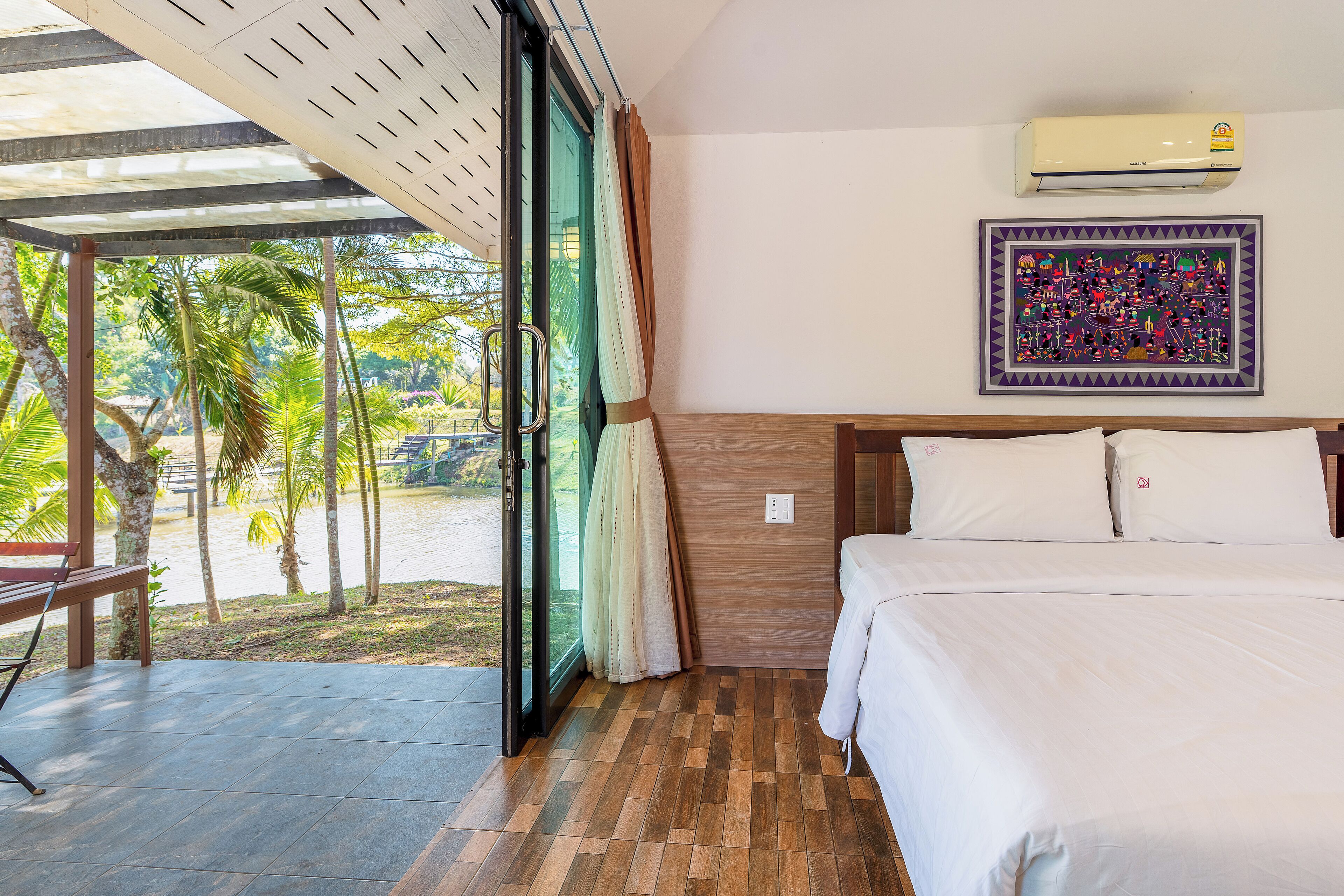 A hotel room with a bed, a window with curtains, an air conditioning unit, and a view of palm trees and water outside.