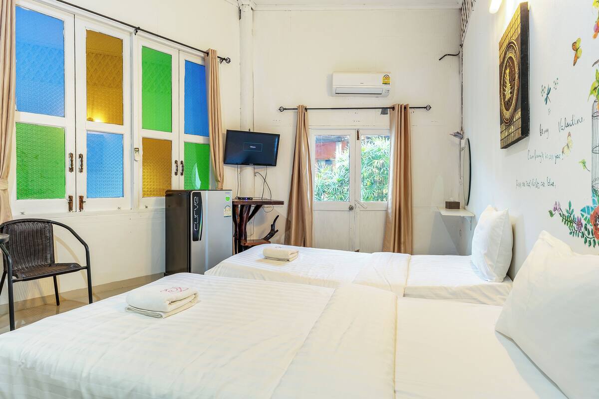 A hotel room with two beds, a television, an air conditioner, and a window with curtains.