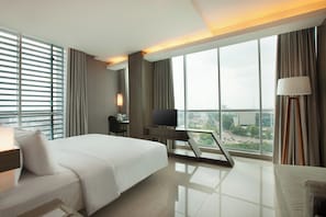 A modern hotel room with a large bed, a desk with a television, a desk lamp, and a view of the city through large windows. - Homm Palembang Radial (Palembang)