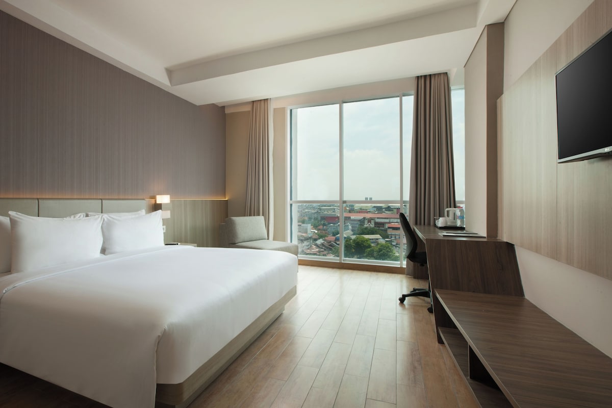 A hotel room with a large bed, a desk, a chair, a flat-screen TV, and a large window with a city view.