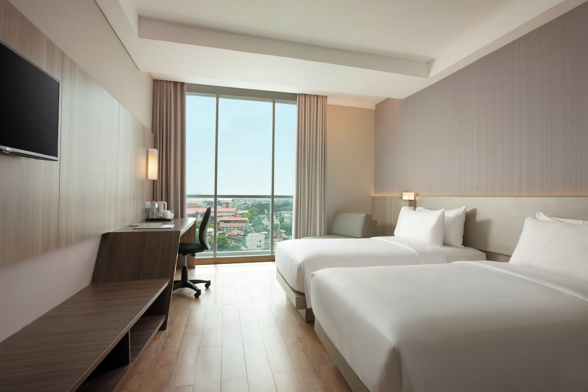 A hotel room with two beds, a desk, a chair, a TV, and a large window offering a city view.