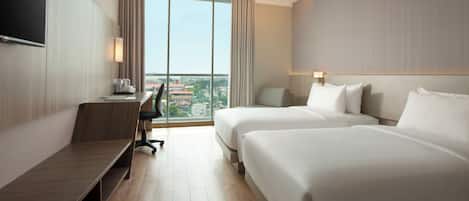 A hotel room with a desk, two beds, a TV, and a large window with a city view.