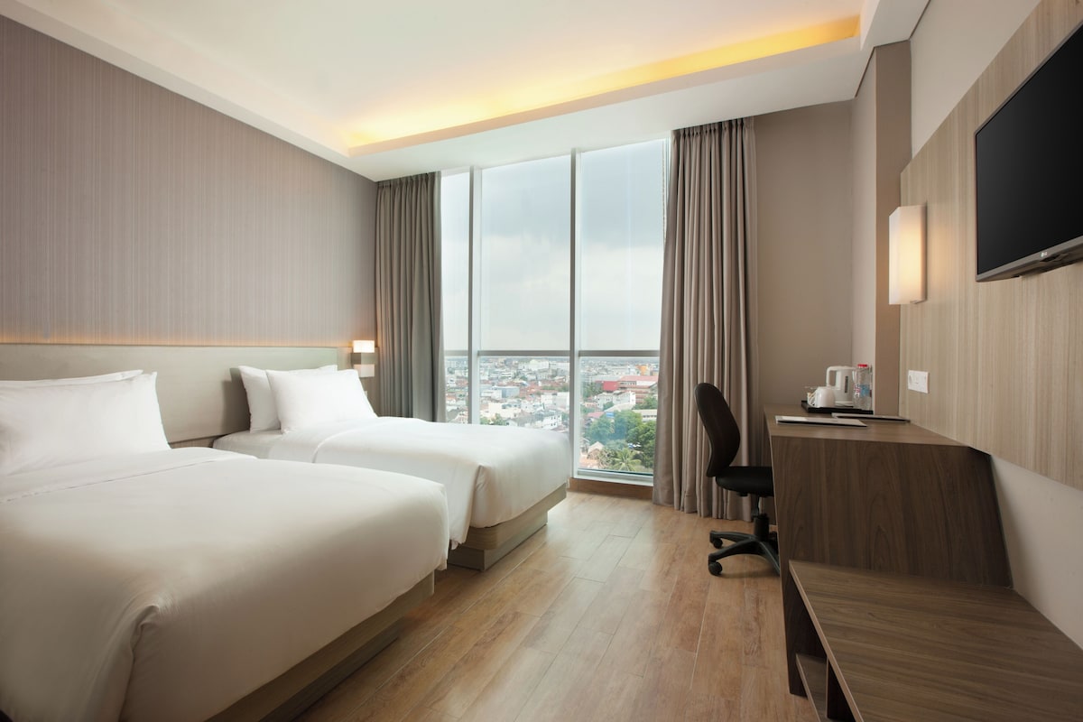 A hotel room with two beds, a desk, a chair, a TV, and a large window with a city view.