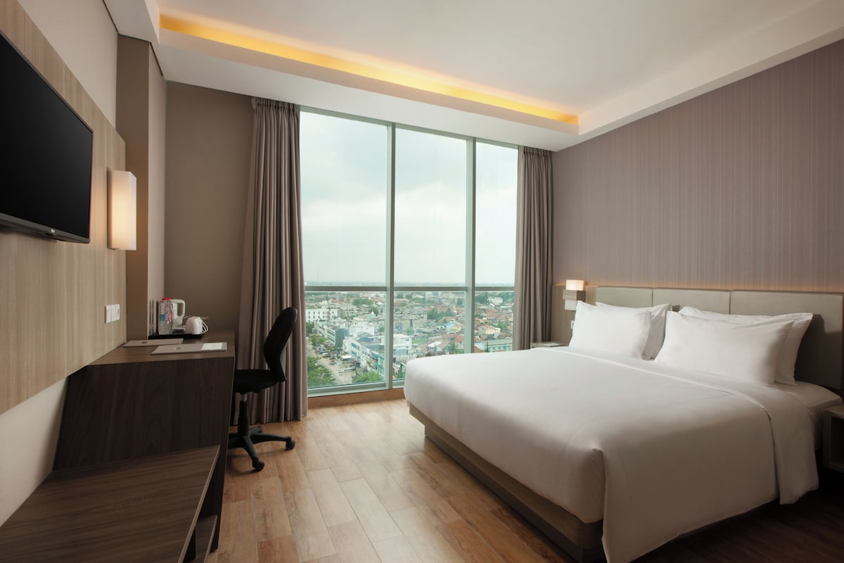 A modern hotel room with a large bed, a desk with a chair, a flat-screen TV, and a large window offering a city view.