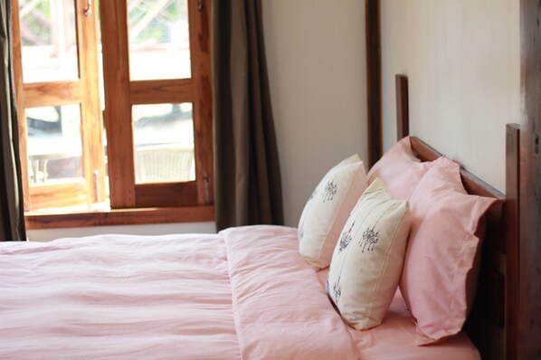 Una cama con ropa de cama rosa y almohadas florales, un cabecero de madera y vista a un balcón a través de una ventana con cortinas.