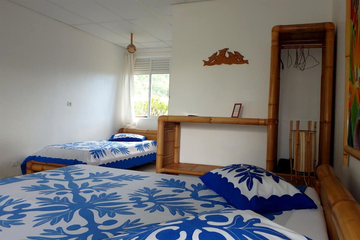 Quarto com duas camas, janela com cortinas, cabeceira de madeira, espelho fixado na parede e uma pintura abstrata colorida.