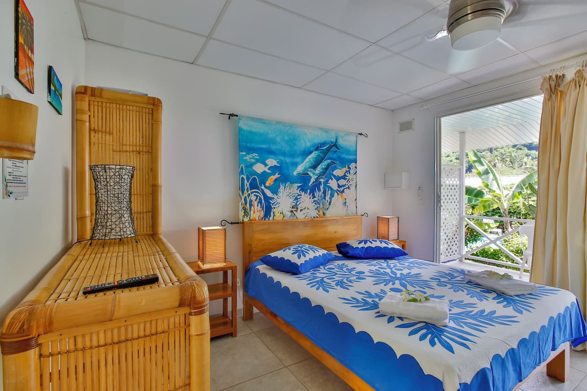 Quarto com cama, uma cadeira de madeira, um quadro e janela com vista para o verde.
