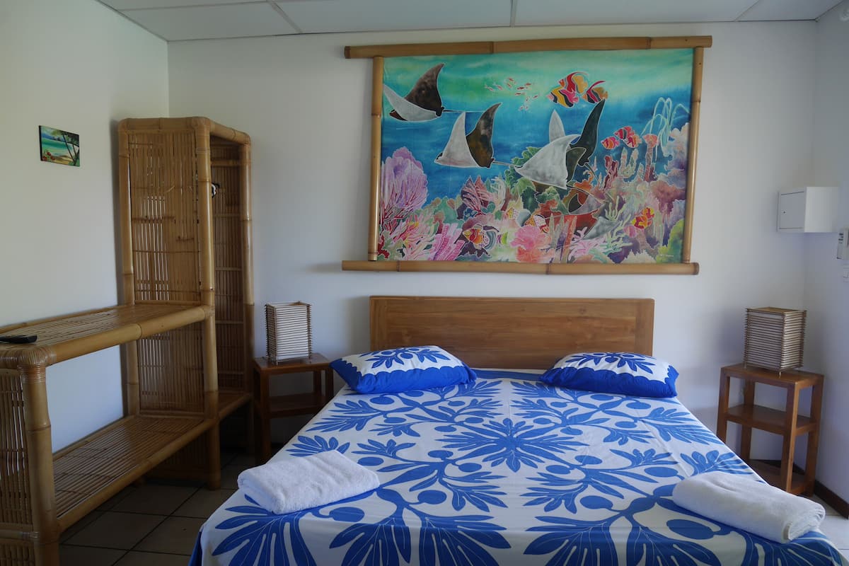 Quarto de hotel com uma cama, duas mesinhas de cabeceira, uma cadeira de madeira, um quadro na parede e uma estante.