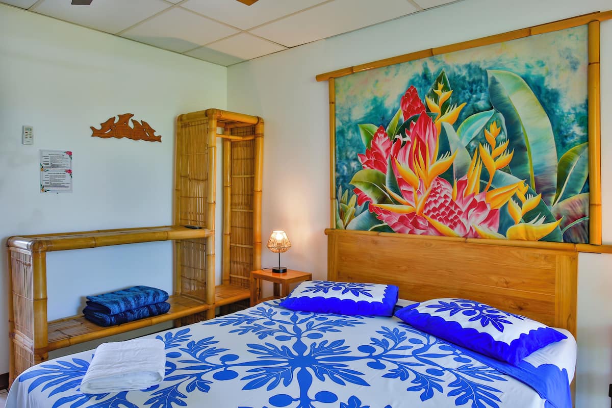 Quarto com cama, cabeceira de madeira, pintura de flores tropicais, mesinha com abajur e cadeira de bambu.