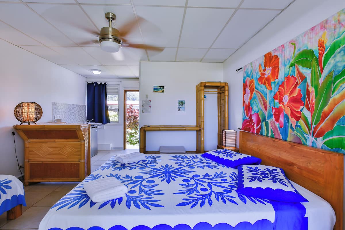 Quarto de hotel com cama, cabeceira de madeira, ventoinha no teto, quadro na parede e pequena mesa com luminária.