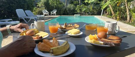 A breakfast table with plates of toast, eggs, fruit, and juice, set against a poolside view with lounge chairs and tropical plants.