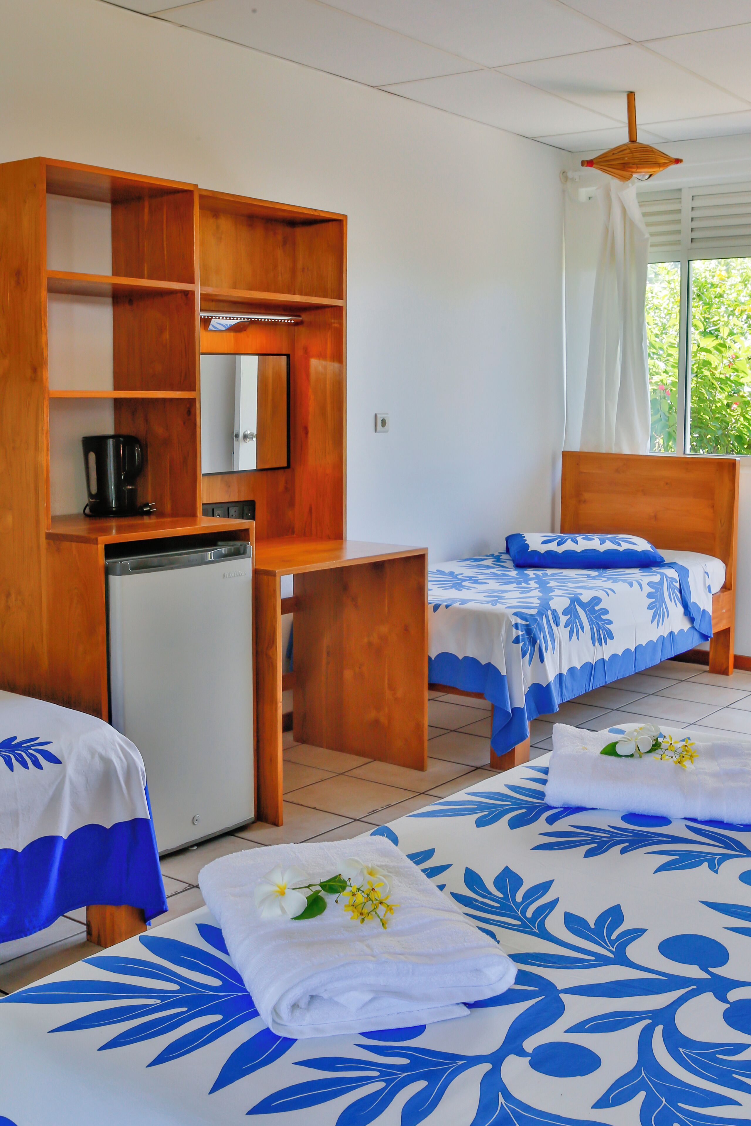 A room with two beds, a small kitchenette, and a window with curtains.
