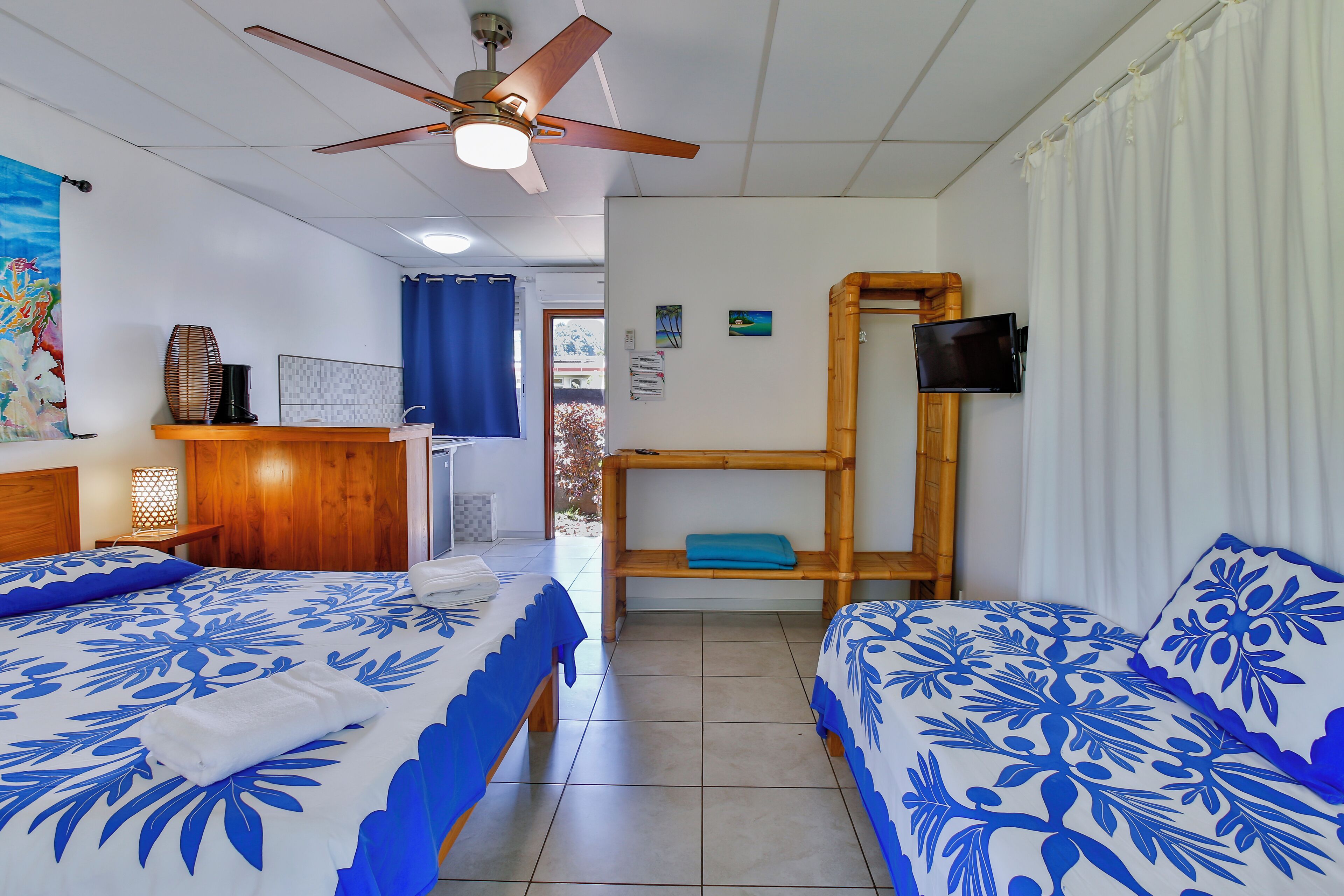 Une chambre avec deux lits, un ventilateur de plafond, un téléviseur fixé sur un cadre en bois et une vue sur l’extérieur par une fenêtre ornée de rideaux bleus.