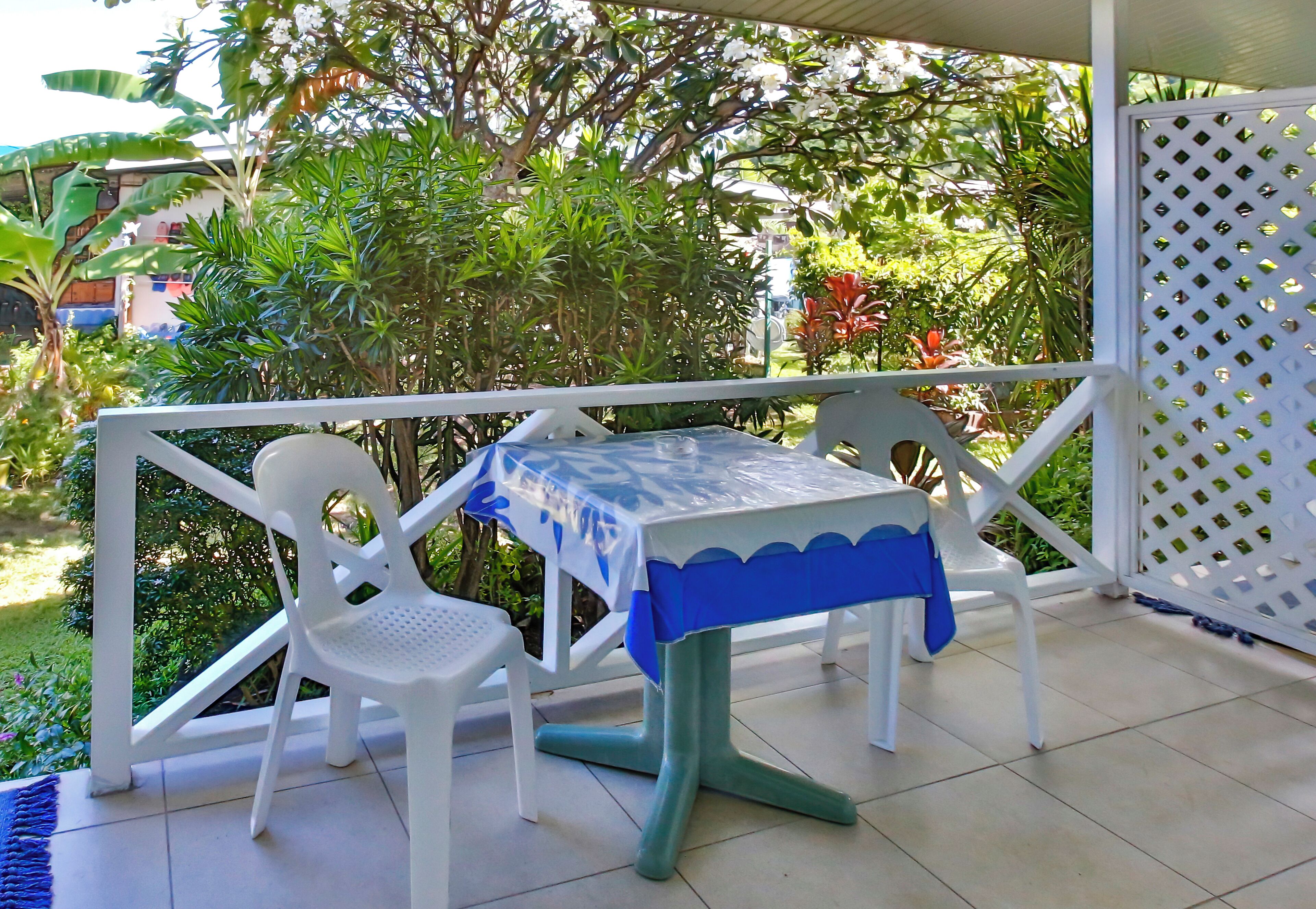A covered patio area with a table and chairs, surrounded by a white lattice fence and lush greenery.