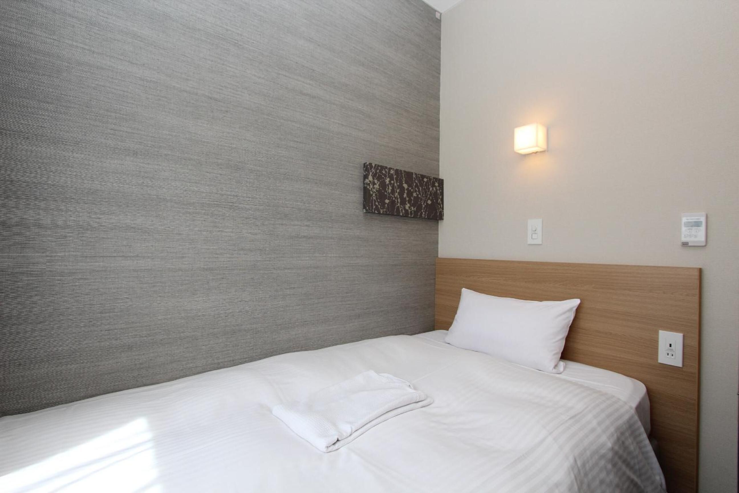 A hotel room with a large bed, a wooden headboard, a textured grey wall, a small artwork, and a wall-mounted light fixture.