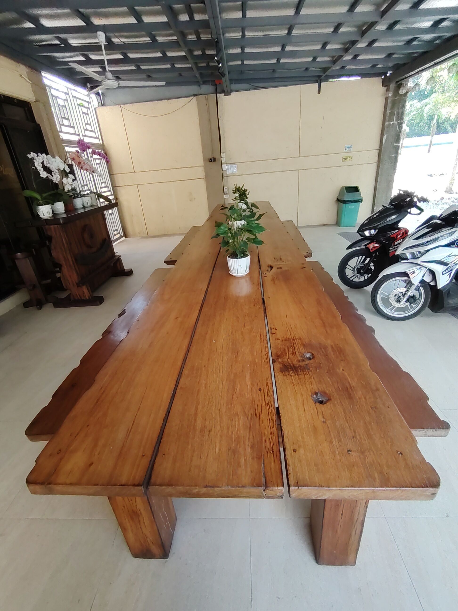 Une longue table en bois, avec un pot de plante placé au centre, un ventilateur de plafond et une moto garée à l’extérieur.