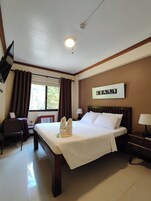 Habitación de hotel con cama, escritorio, silla, televisor, ventana con cortinas y una obra de arte colgada en la pared.