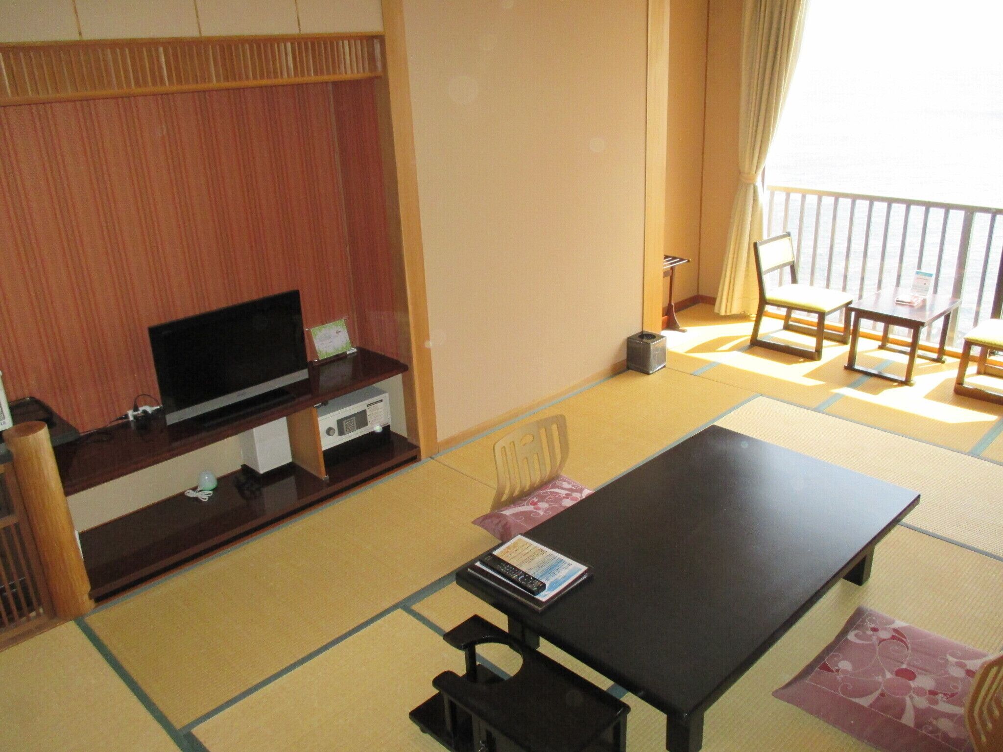 Una habitación japonesa tradicional con suelo de tatami, una mesa de madera baja, un televisor y una puerta corredera que da a un balcón.