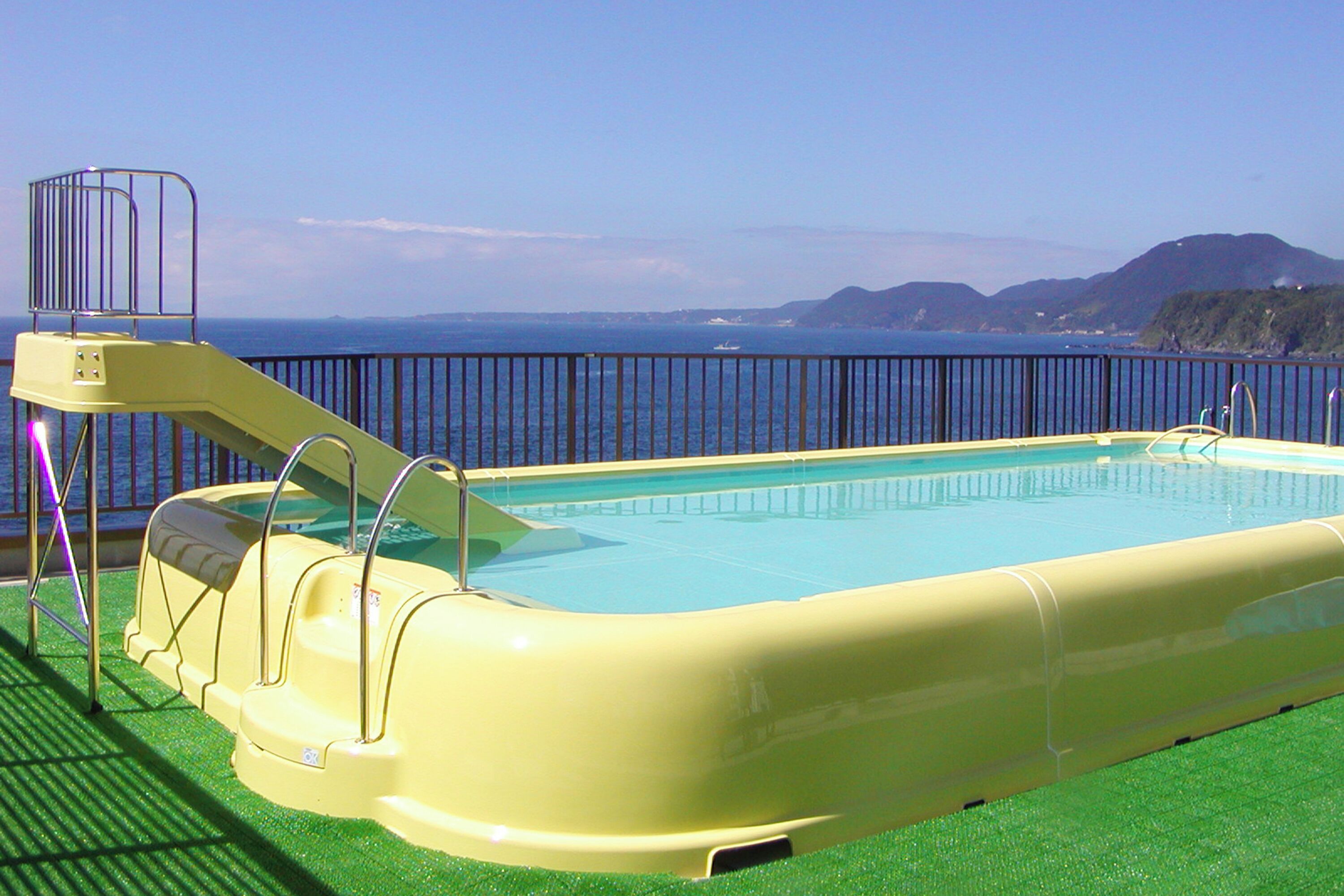 Una piscina con tobogán, rodeada por una barandilla, con vistas al mar y colinas al fondo.