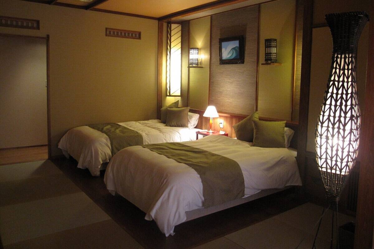 A hotel room with two beds, a nightstand with a lamp, a wall-mounted TV, and a decorative hanging light fixture.