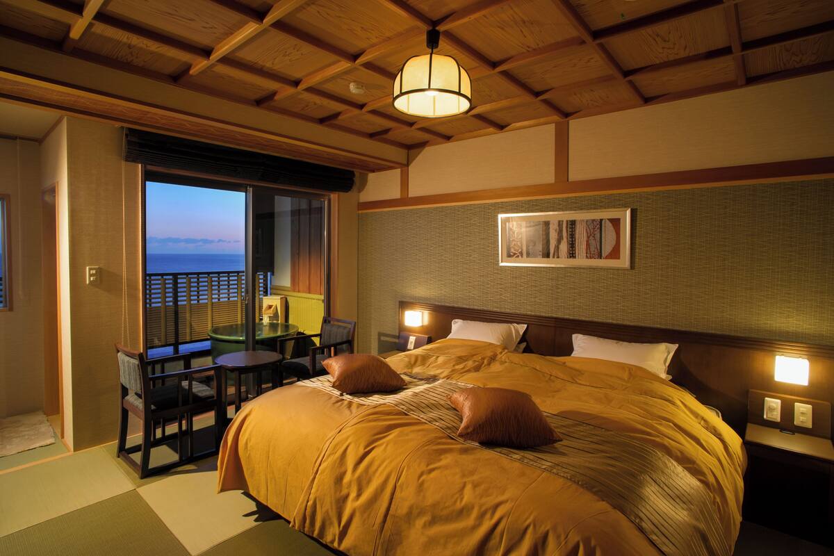 A hotel room with a large bed, a desk with a chair, a view of the sea, and a ceiling with wooden beams.