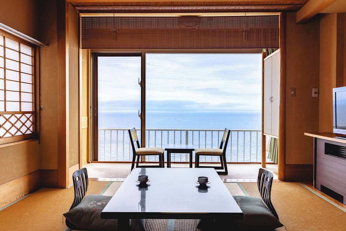 A room with a view of the sea, a table with chairs, and a TV.