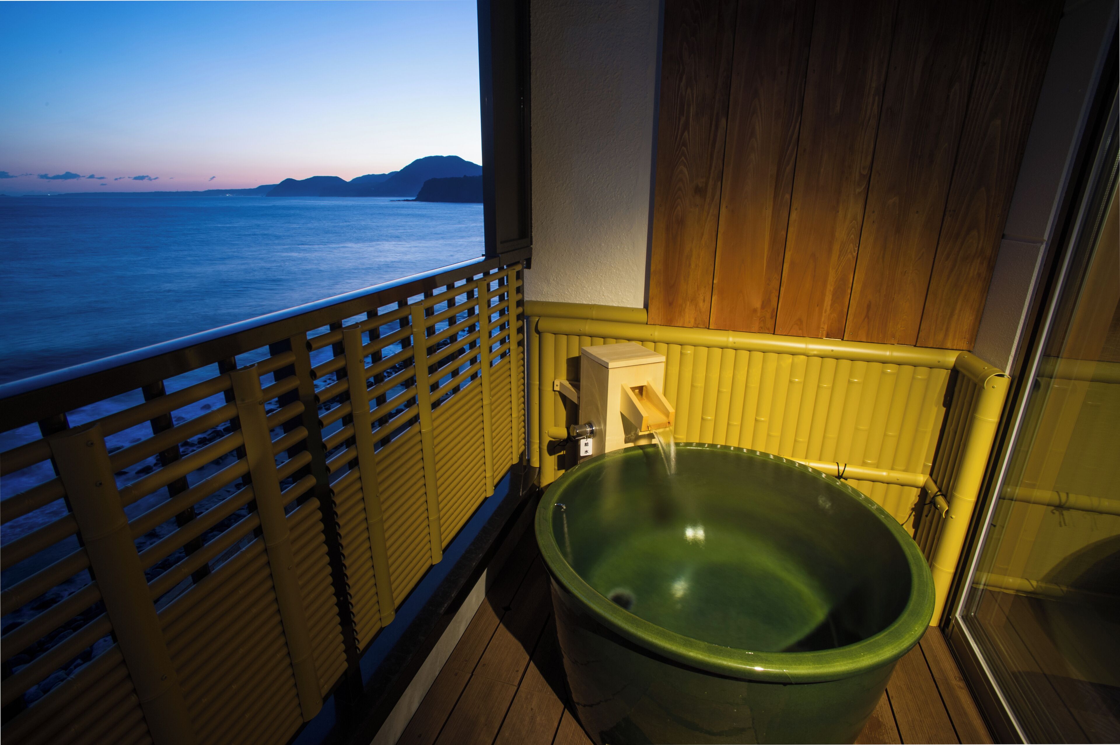 A balcony with a view of the sea, a wooden floor, and a green bathtub.