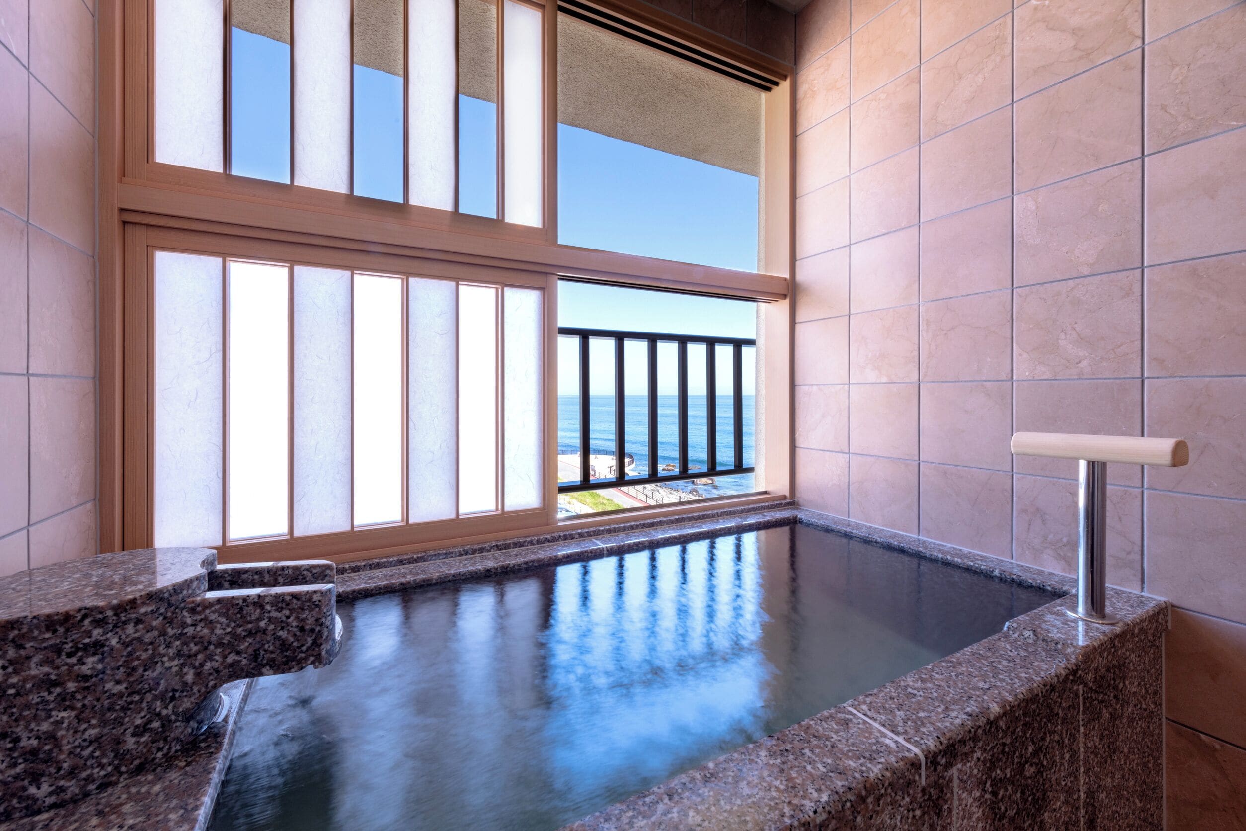 Ein Wellnessbereich mit Whirlpool, Meerblick durch große Fenster und ein Balkon mit Geländer.