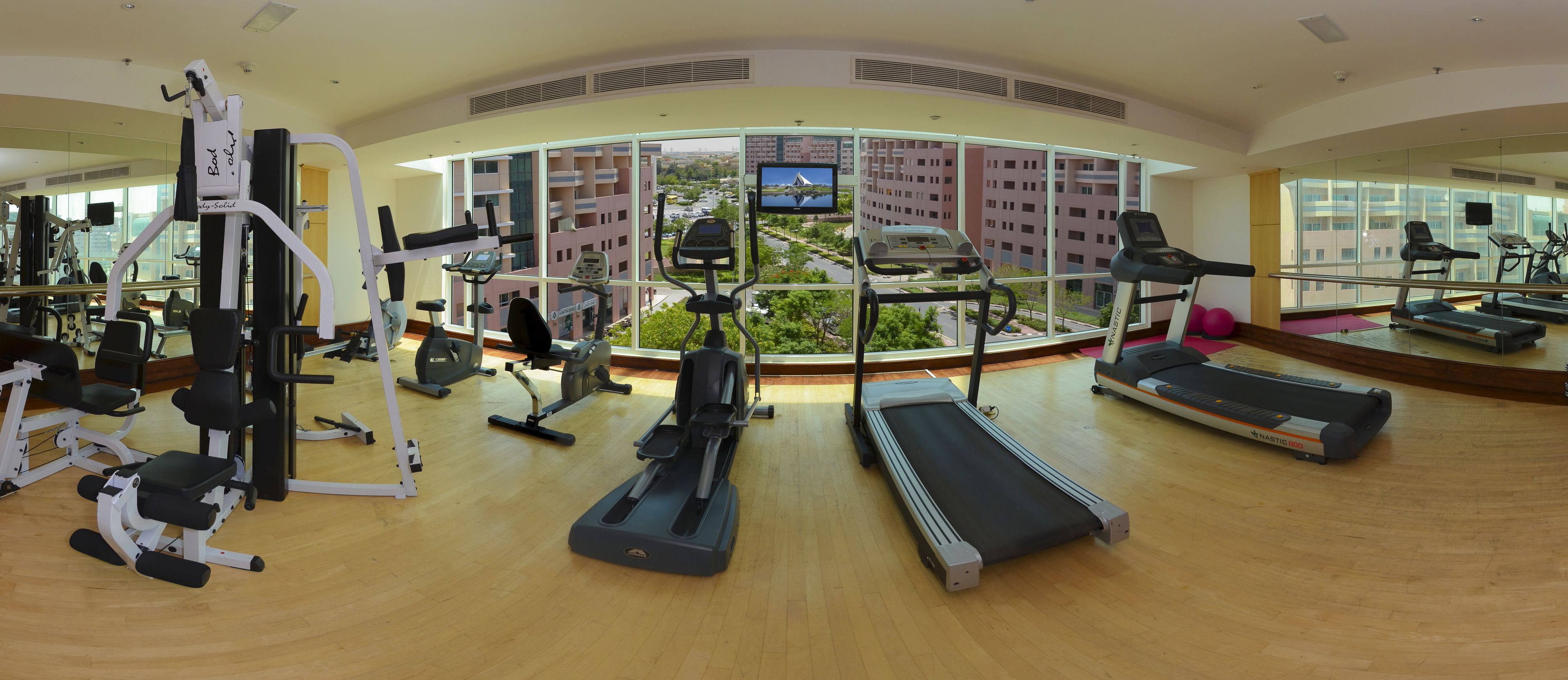 A gym with treadmills, exercise bikes, and weight machines, featuring large windows with a city view.