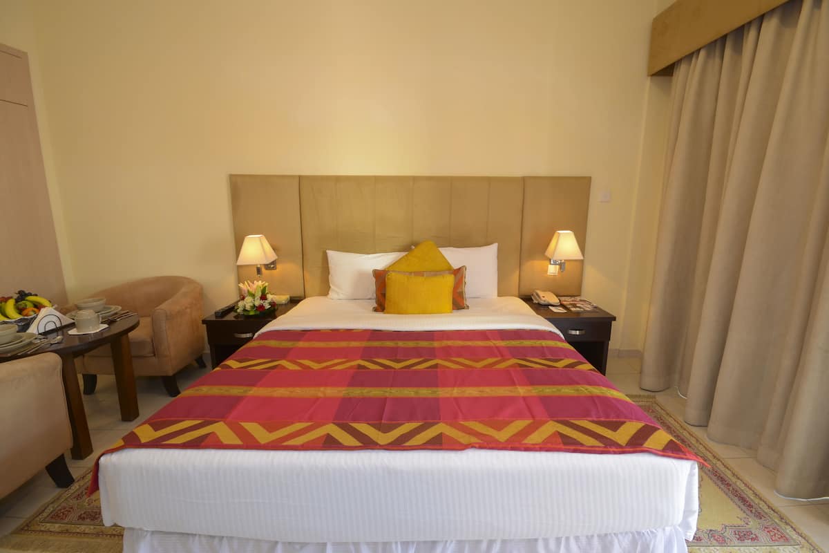 Une chambre d’hôtel avec un grand lit, deux tables de chevet avec des lampes, une chaise et une petite table sur laquelle se trouvent un panier de fruits et des tasses.
