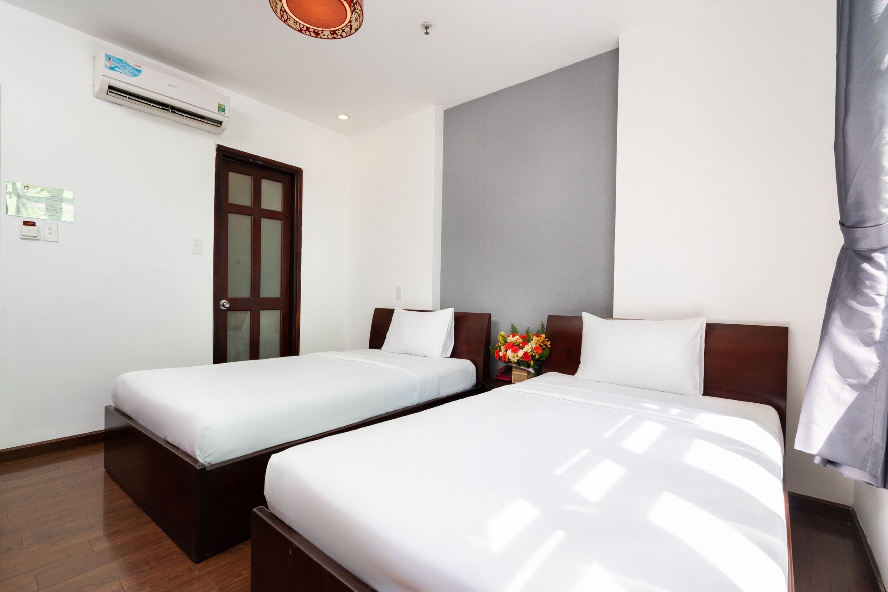 A hotel room with two single beds, a wooden floor, a large grey wall, and an air conditioning unit.