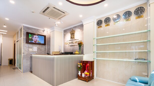 A hotel reception area with a reception desk, a TV screen, clocks displaying times from various cities, and a blue sofa. -  A&A Toronto Hotel (Ho Chi Minh City)