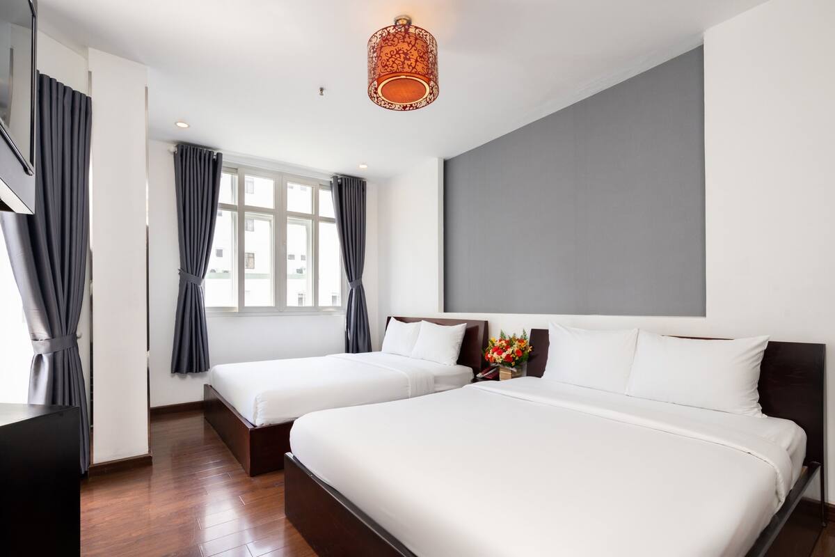 Een hotelkamer met twee bedden, een groot raam, grijze gordijnen, een houten vloer en een moderne lamp.
