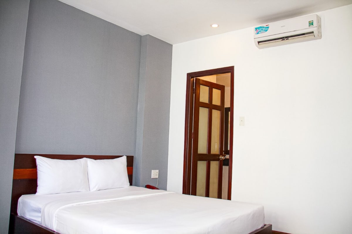 A hotel room with a bed, a wooden headboard, a white wall, a grey accent wall, a wooden door, and an air conditioning unit.
