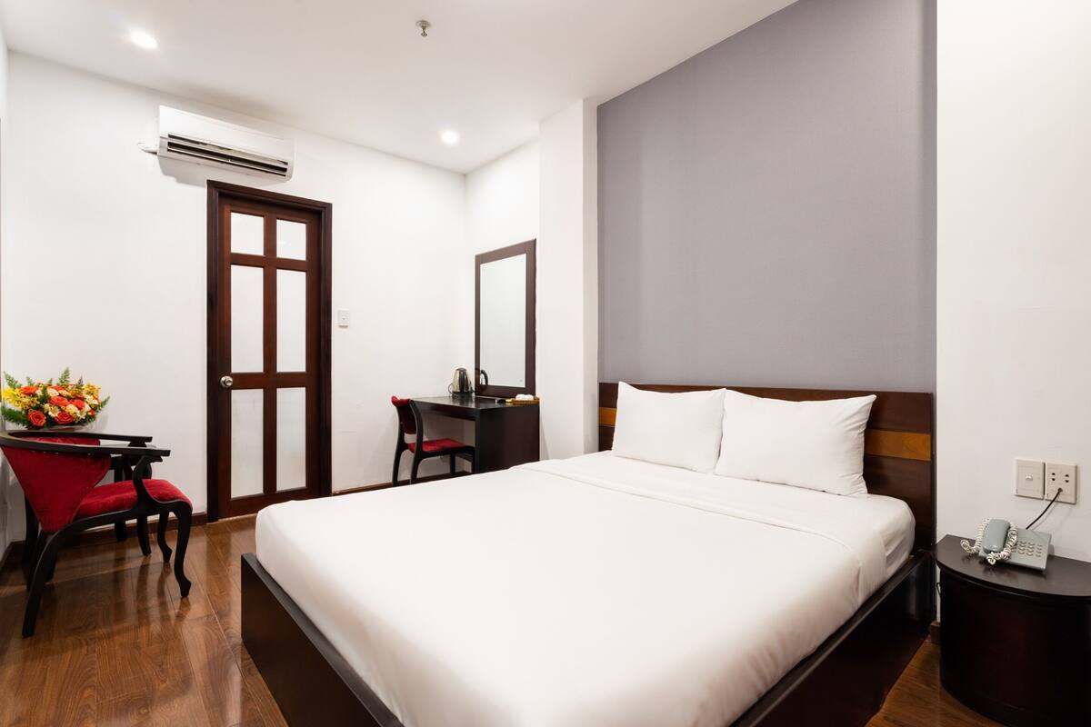 A hotel room with a large bed, a desk with a chair, a mirror, and an air conditioning unit.