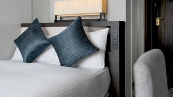 Una cama bien hecha con dos almohadas, una mesita de noche con una lámpara y una puerta a la derecha.