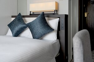 Una cama bien hecha con dos almohadas, una mesita de noche con una lámpara y una puerta a la derecha.