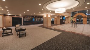 Un vestíbulo amplio con techo alto, iluminación empotrada y un candelabro central. Hay zonas de estar con sillas y una mesita, además de un mostrador de recepción con una pantalla digital.