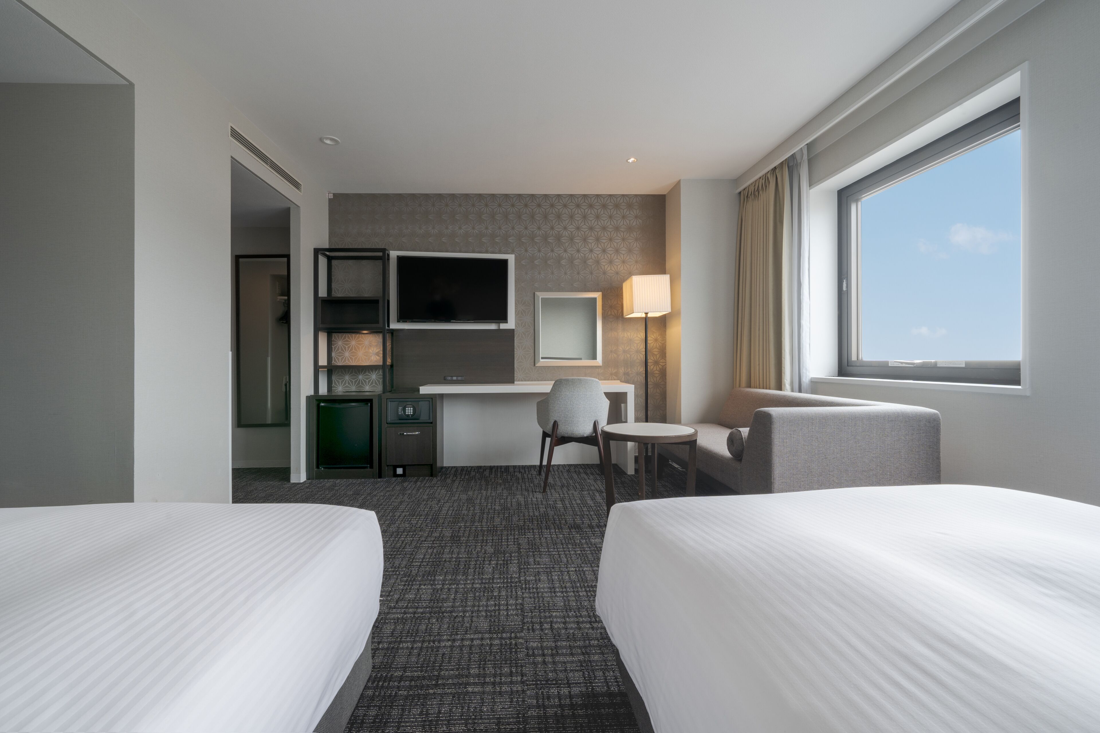 A hotel room with two beds, a desk, a chair, a TV, and a window.