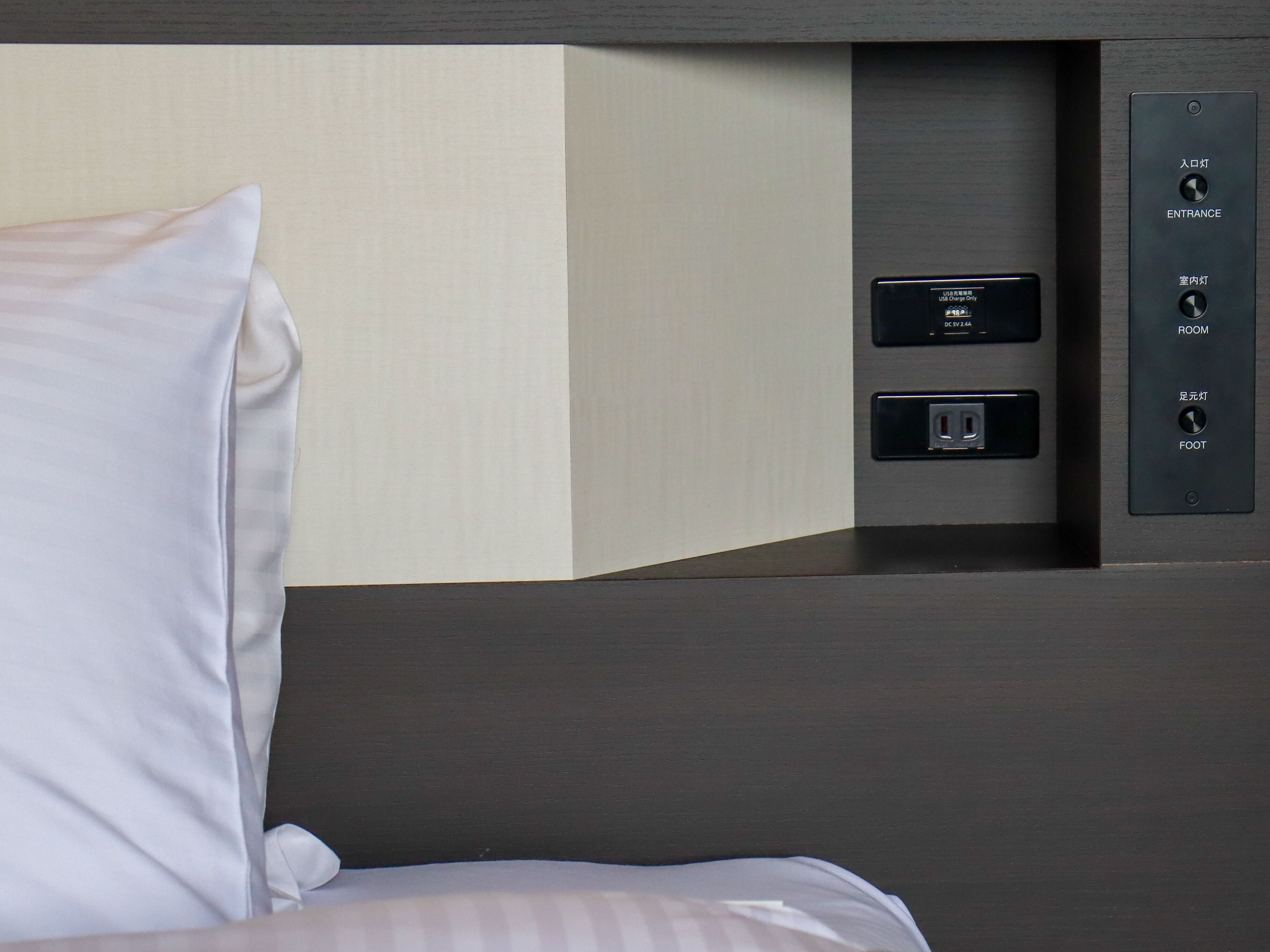 A hotel room with a bed, a control panel with buttons labeled "Entrance," "Room," and "Foot," and a USB charging port.