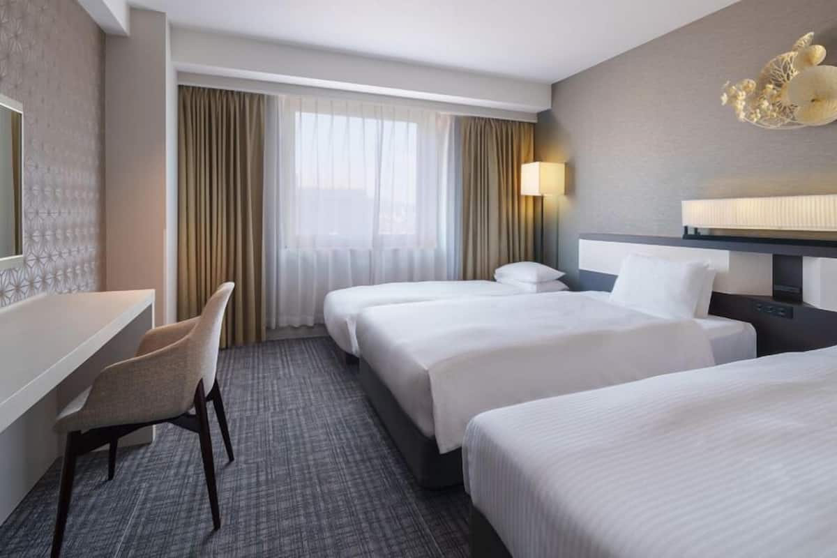 A hotel room with a desk, two beds, a chair, a lamp, and a decorative wall piece.
