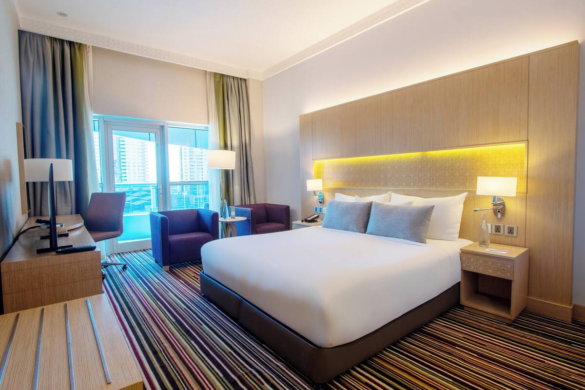 A hotel room with a large bed, two bedside tables with lamps, a desk with a chair, a small table, and a view of the city through large windows.