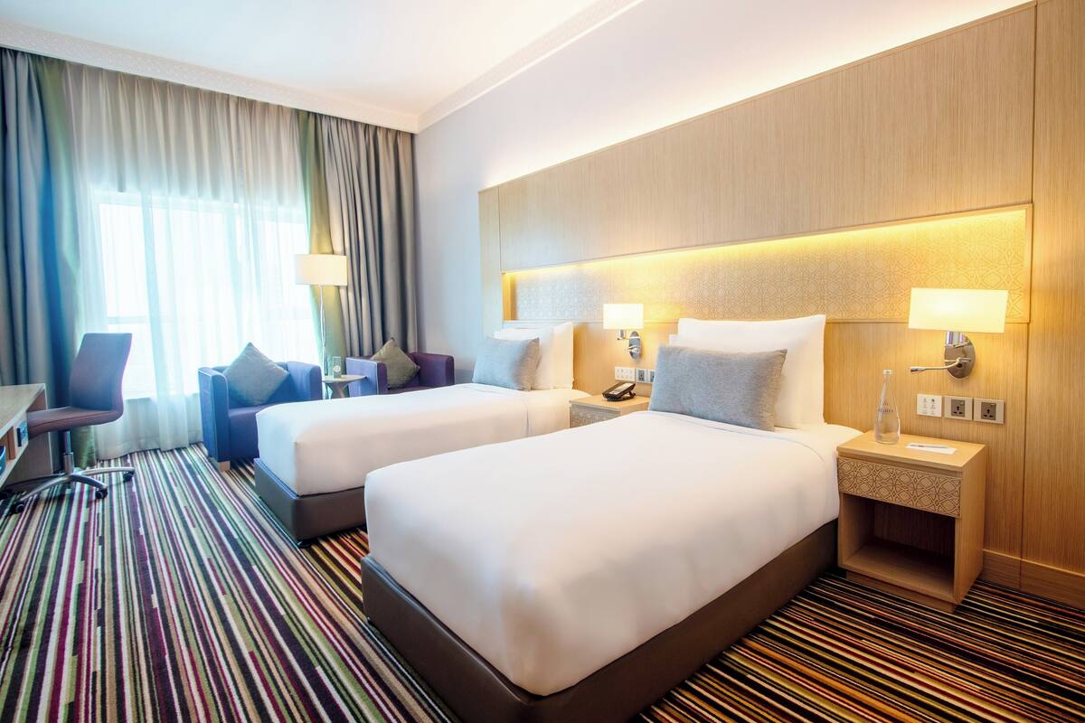 A hotel room with two beds, a desk, a chair, and a striped carpet.