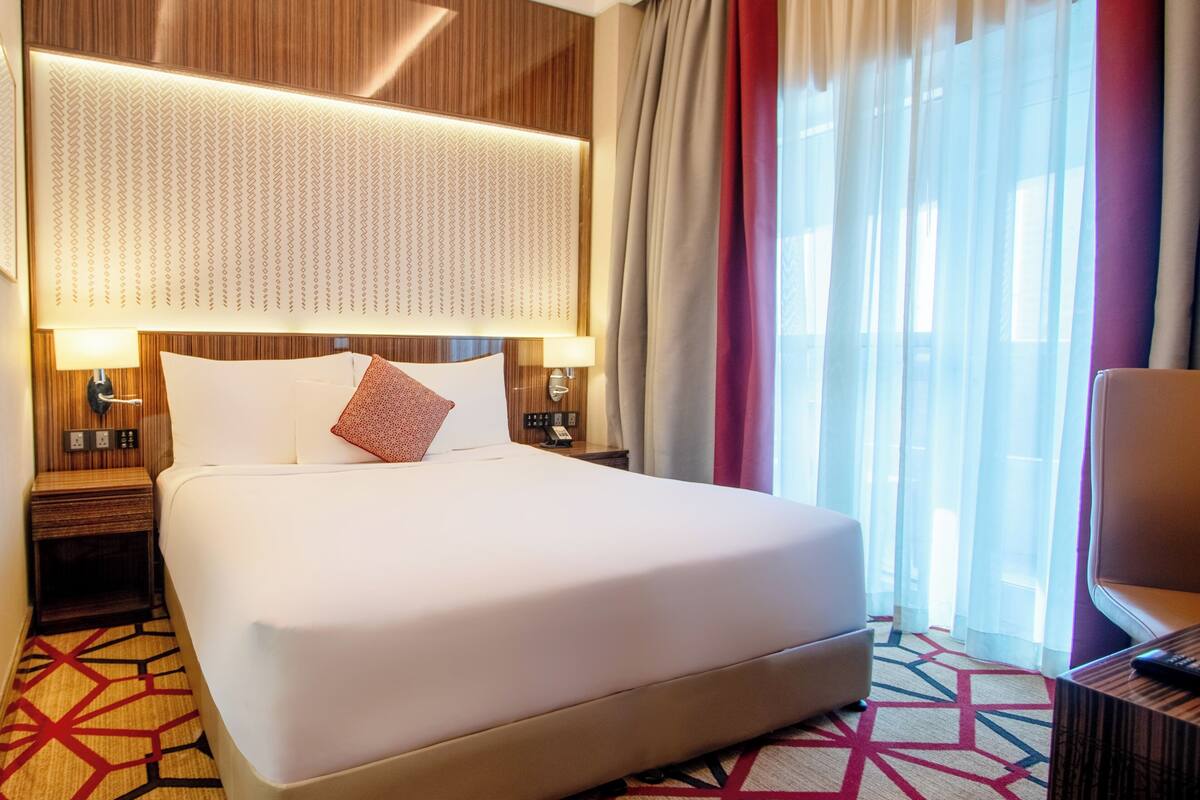 A hotel room with a large bed, a wooden headboard, a patterned wall, a nightstand with a lamp, and a view of the outdoors through sheer curtains.