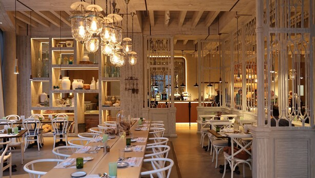 Un restaurante con largas mesas de madera y sillas blancas, candelabros de cristal colgantes, y estantes con cristalería y vajilla.