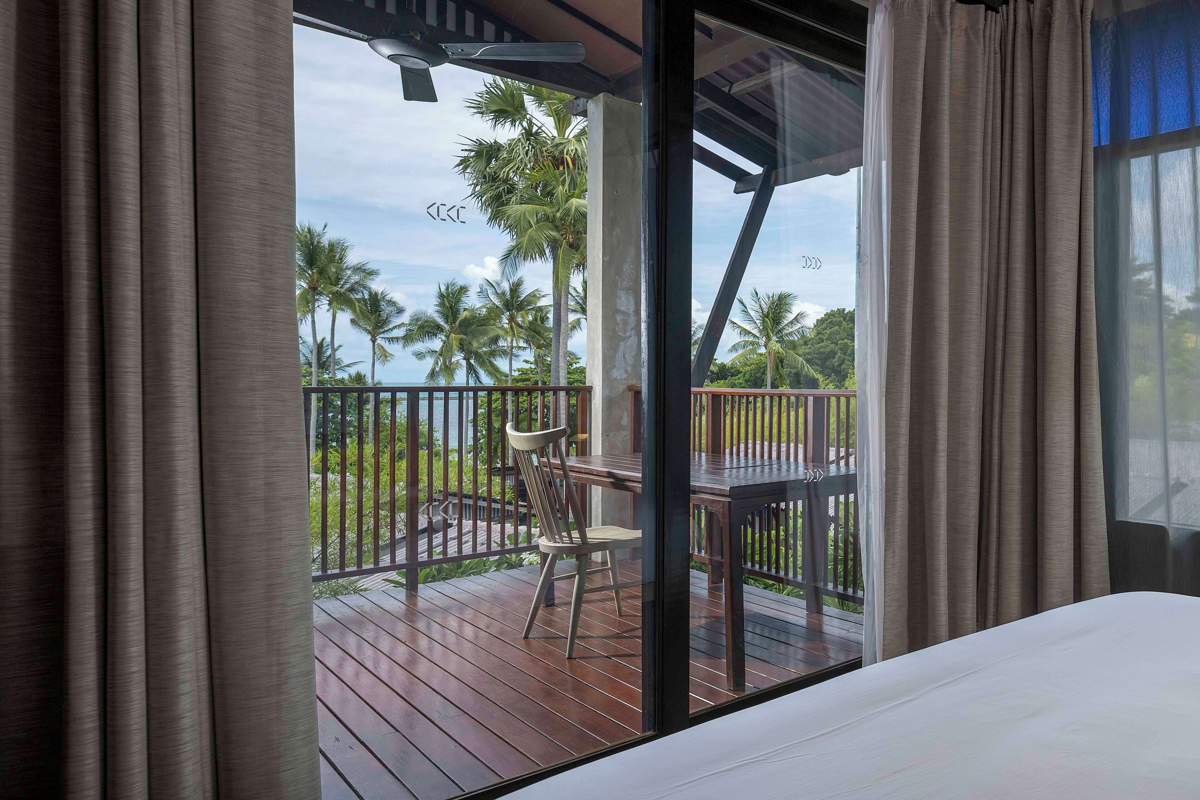 A room with a bed, a wooden deck, a chair, and a view of palm trees and the sea.