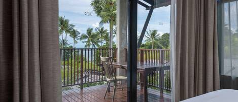 A room with a bed, a wooden deck, a chair, and a view of palm trees and the sea.