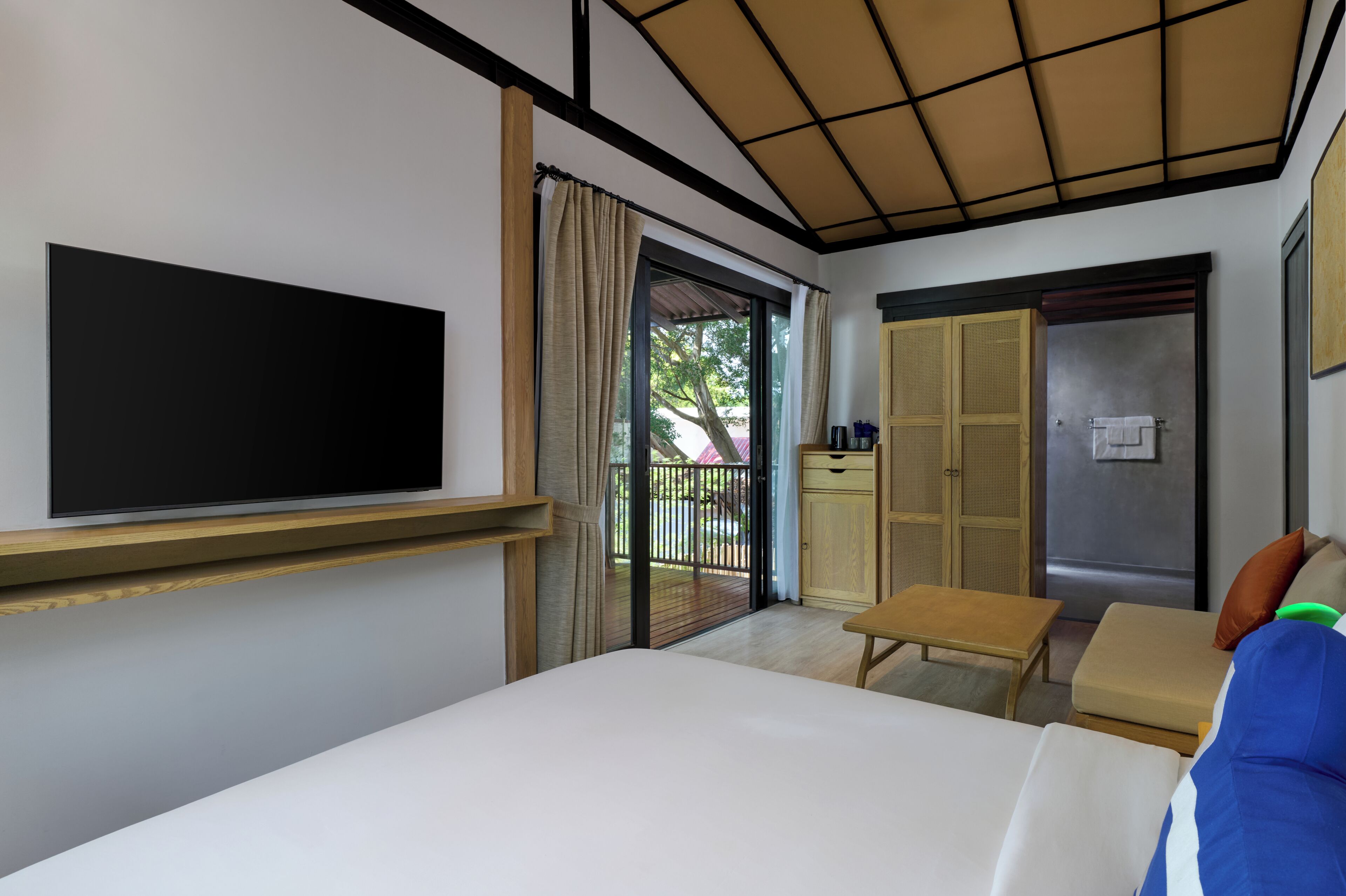 A hotel room with a bed, a TV mounted on the wall, a sliding glass door to the balcony, a small table, and a wardrobe.